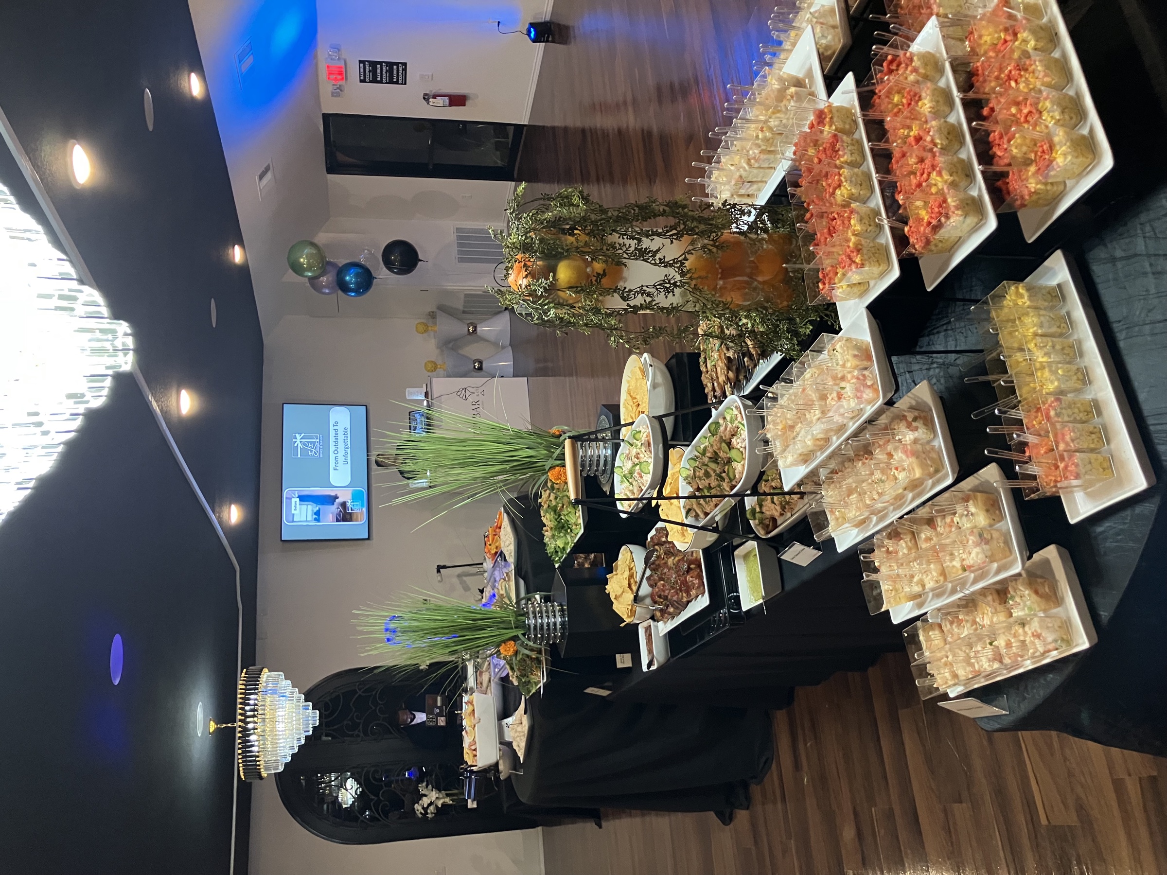 A lavish corporate appetizer buffet with chafing dishes, sushi cups, and decorative greenery, lit by chandeliers and blue accent lighting.